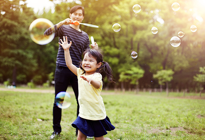 father blowing bubbles and young girl chasing them in a park on a summer day